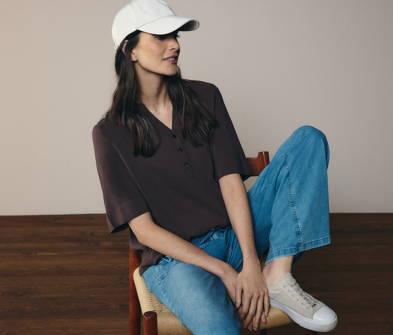 Žena sedí na stoličke, má džínsy, šiltovku a hnedú tunikovú blúzku CECIL.