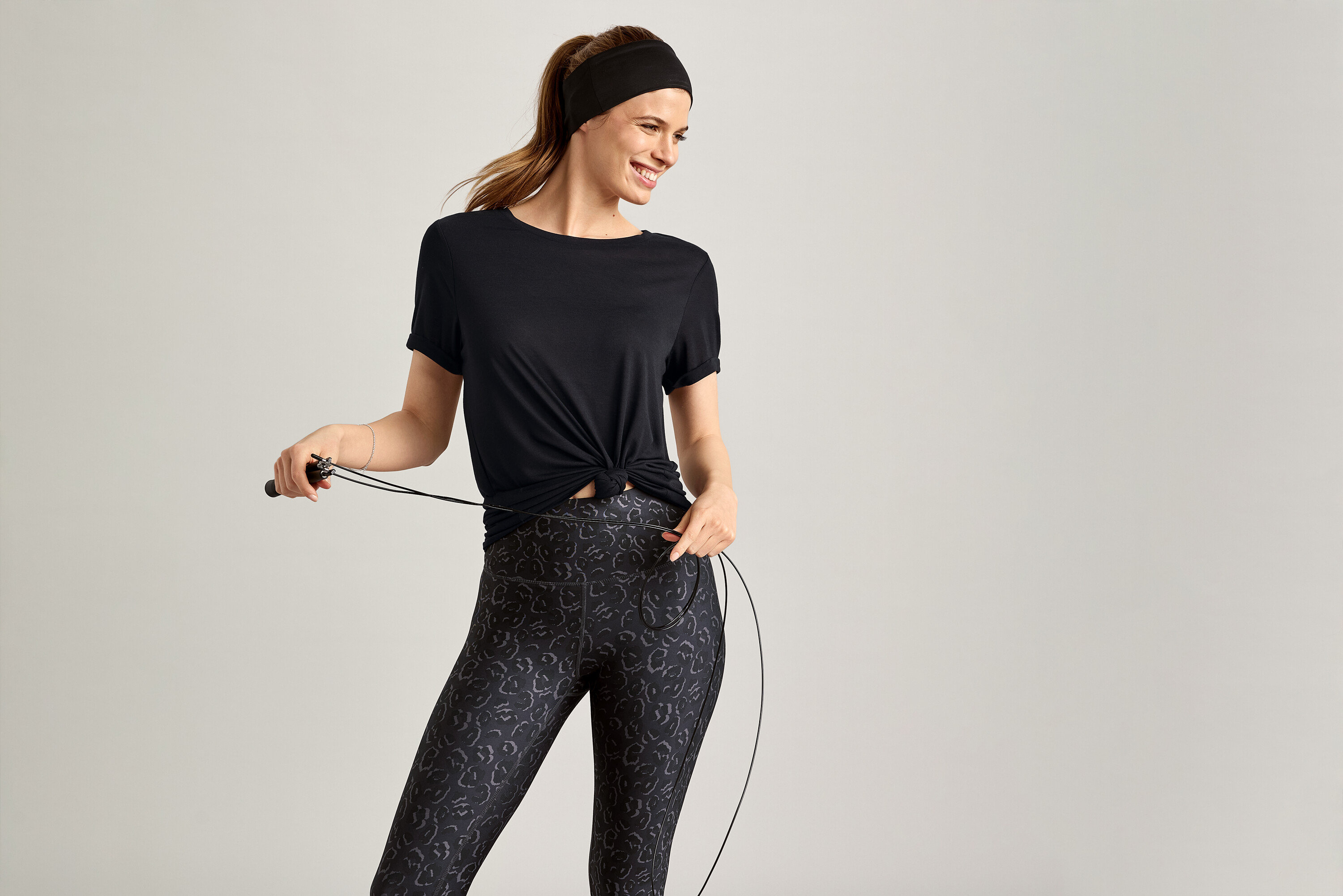 Žena s čelenkou a v čiernom športovom tričku drží švihadlo na sivom pozadí.