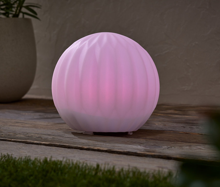 Ružová guľová lampa stojí na drevenom stole vedľa kvetináča s rastlinou.
