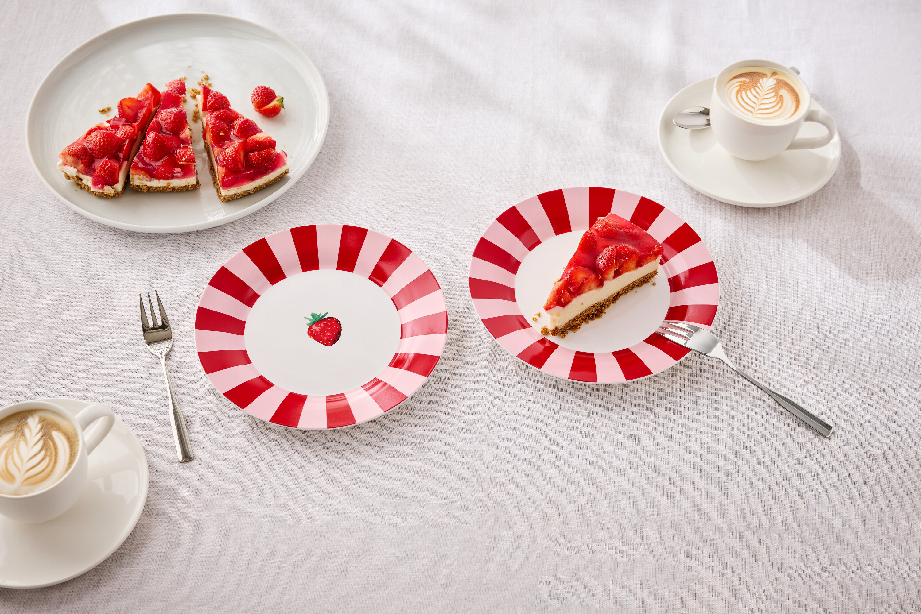 Na stole sú taniere s jahodovým koláčom, vidličky a šálky s latte art.