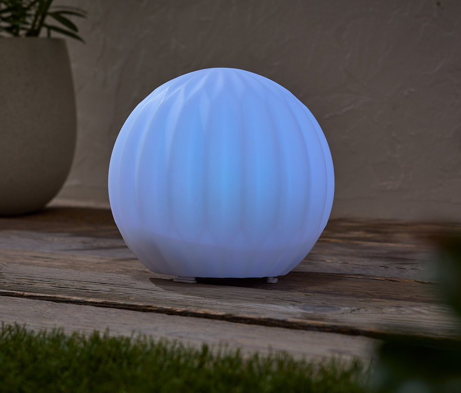 Svietiaca guľovitá lampa stojí na drevenom stole vedľa kvetináča s rastlinou.