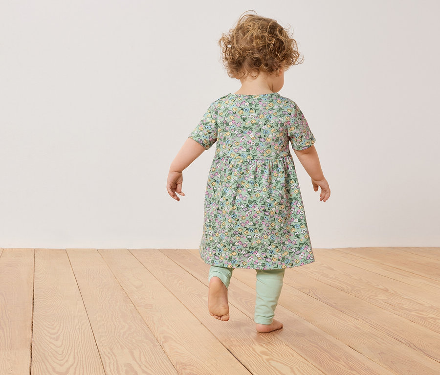 Malé dieťa s kučeravými vlasmi kráča naboso v zelených kvetovaných šatách a legínach po drevenej podlahe.