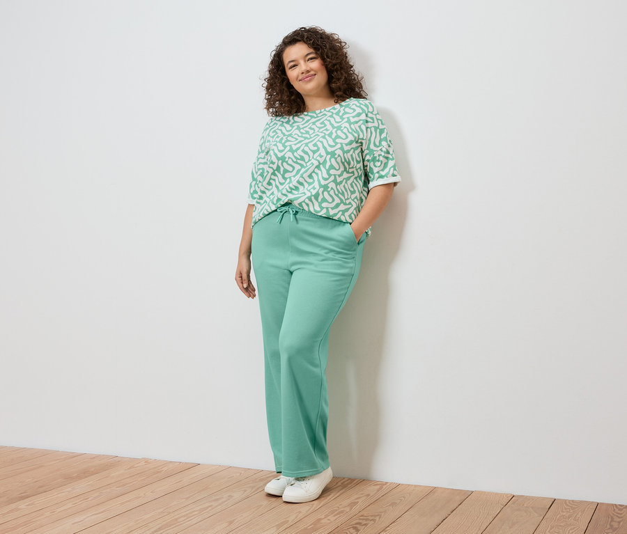 Žena pózuje pri stene, má na sebe svetlozelený vzorovaný top, svetlozelené nohavice a biele tenisky.