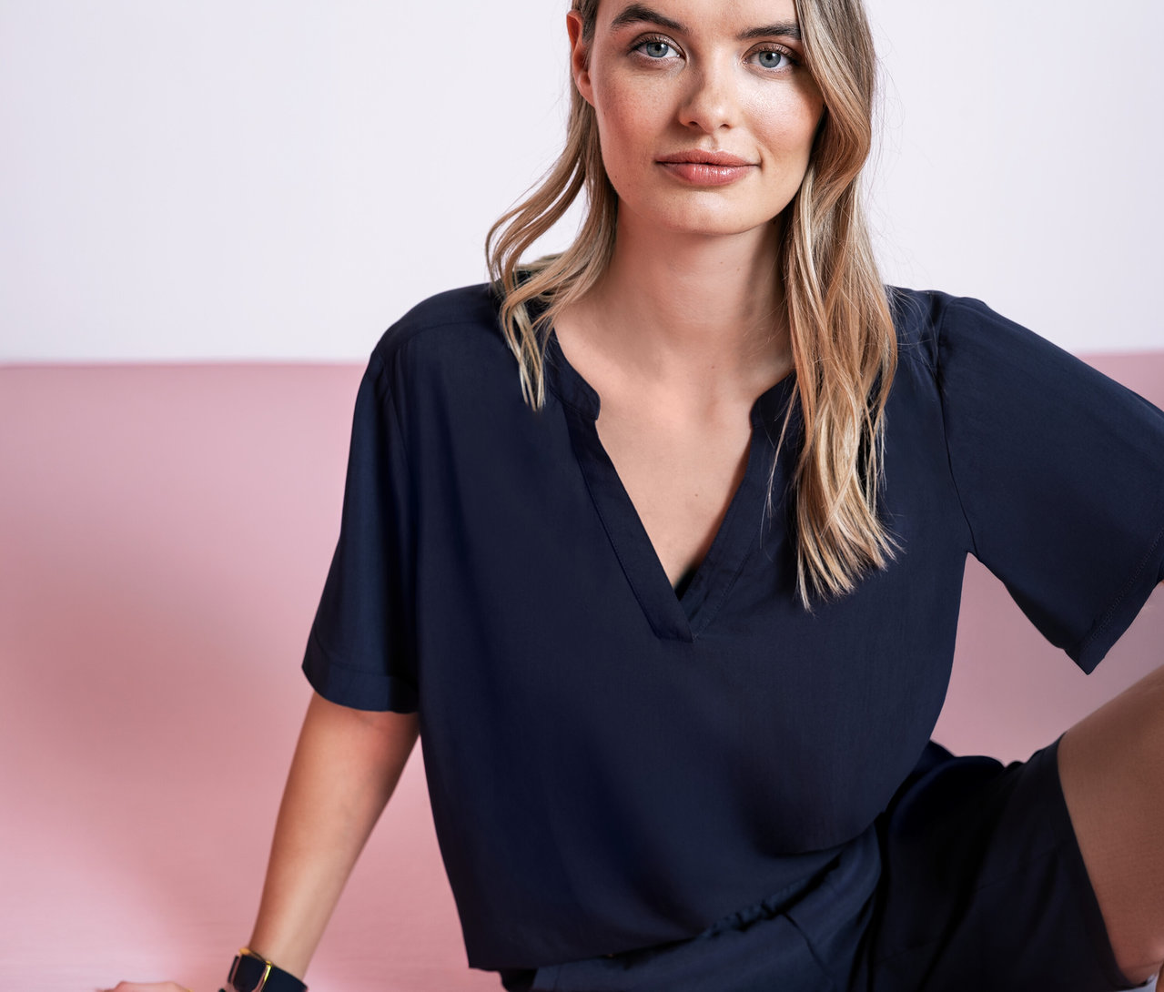 Žena sedí na ružovom pozadí a má na sebe tmavomodrú tkanú blúzku Street One.