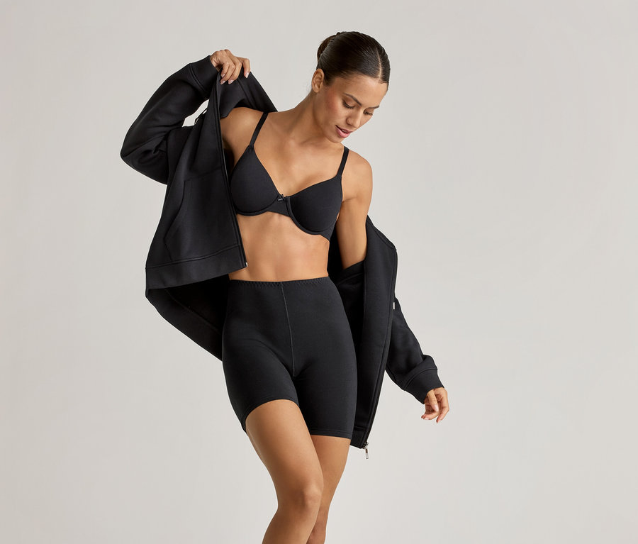 Žena v čiernej podprsenke s kosticami, cyklistických šortkách (2 ks) a pohodlnej mikine, ktorú si rozopína.