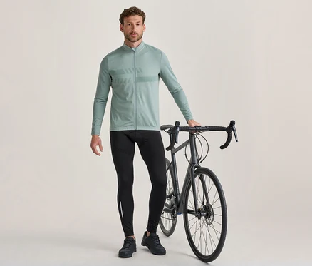 Muž stojí vedľa tmavosivého cestného bicykla a drží jeho riadidlá. Má na sebe svetlozelený cyklistický dres s dlhým rukávom a vzorom na hrudi, čierne cyklistické legíny s reflexným pruhom a čierne cyklistické topánky.