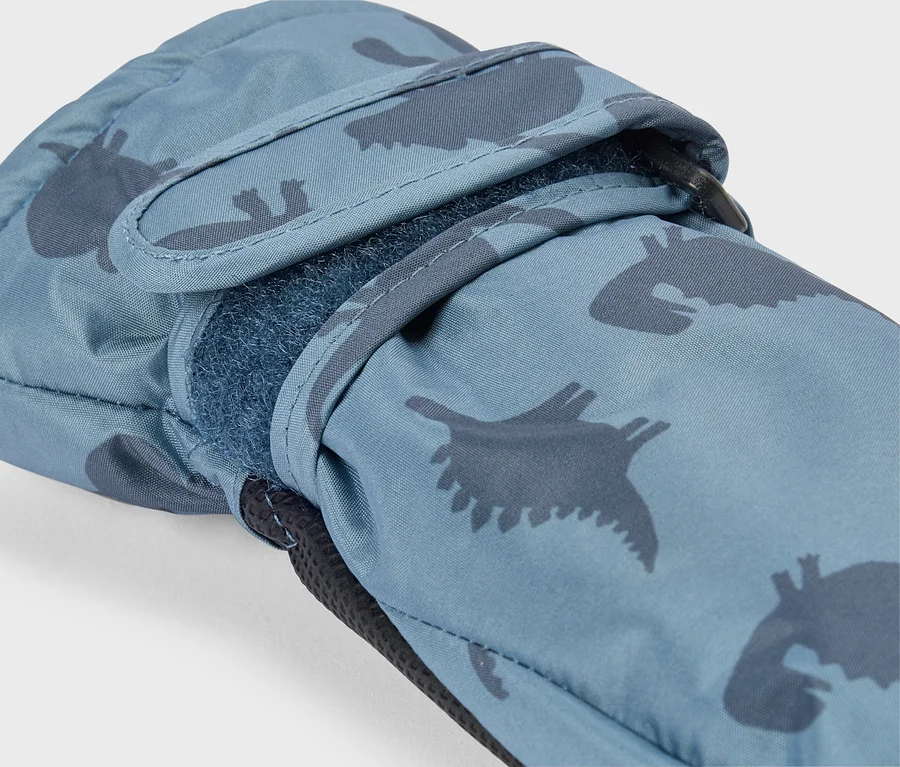 Detail modrej rukavice s tmavomodrým vzorom dinosaurov. Suchý zips na zápästí je čiastočne otvorený, odhaľujúc chlpatú spodnú stranu. Pri zápästí je viditeľná čierna slučka.