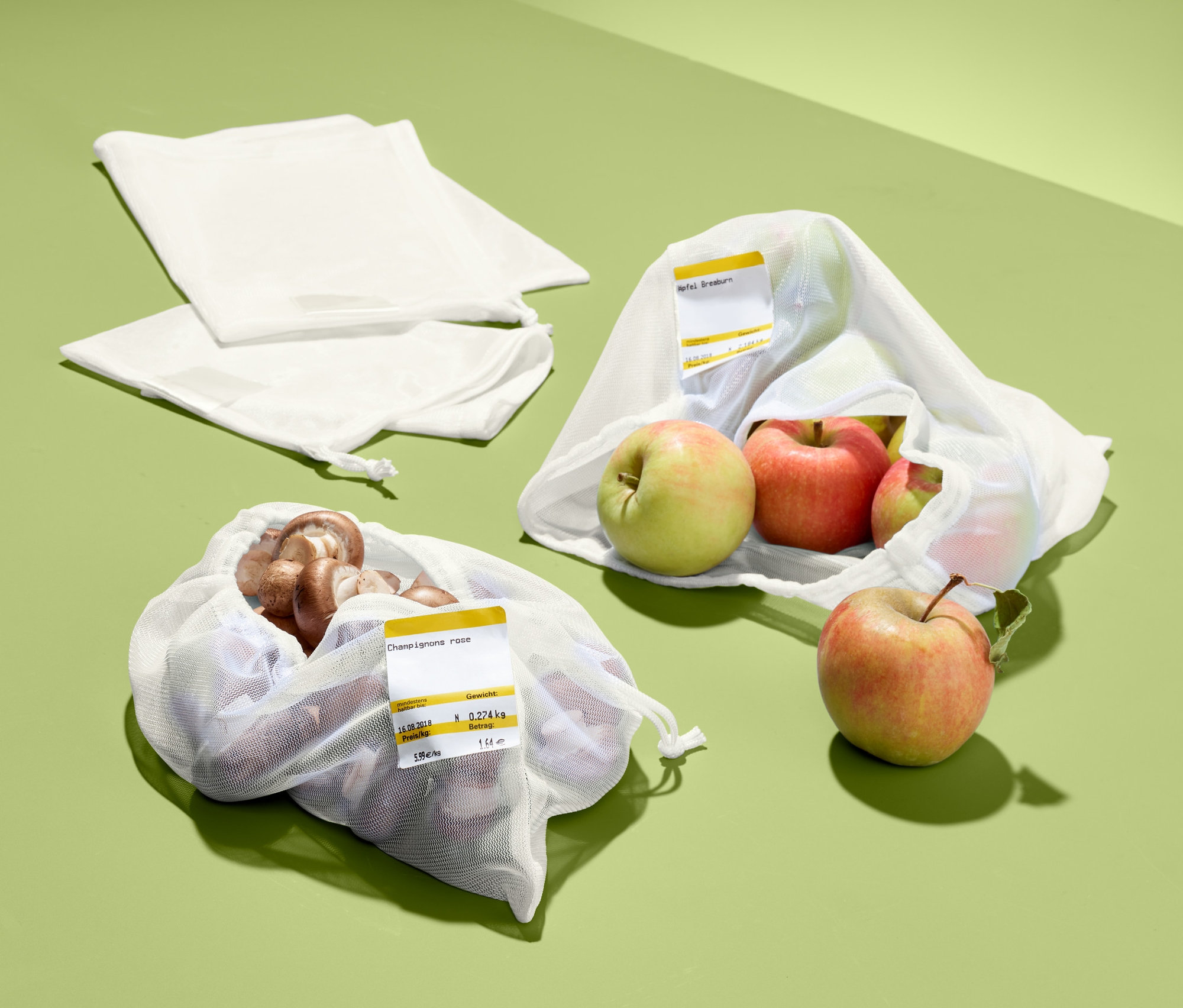 Na zelenom pozadí sú dve voľné biele sieťky a dve naplnené. V jednej sieťke sú jablká a v druhej hnedé huby. Sú to Sieťky na ovocie a zeleninu, 4 ks.