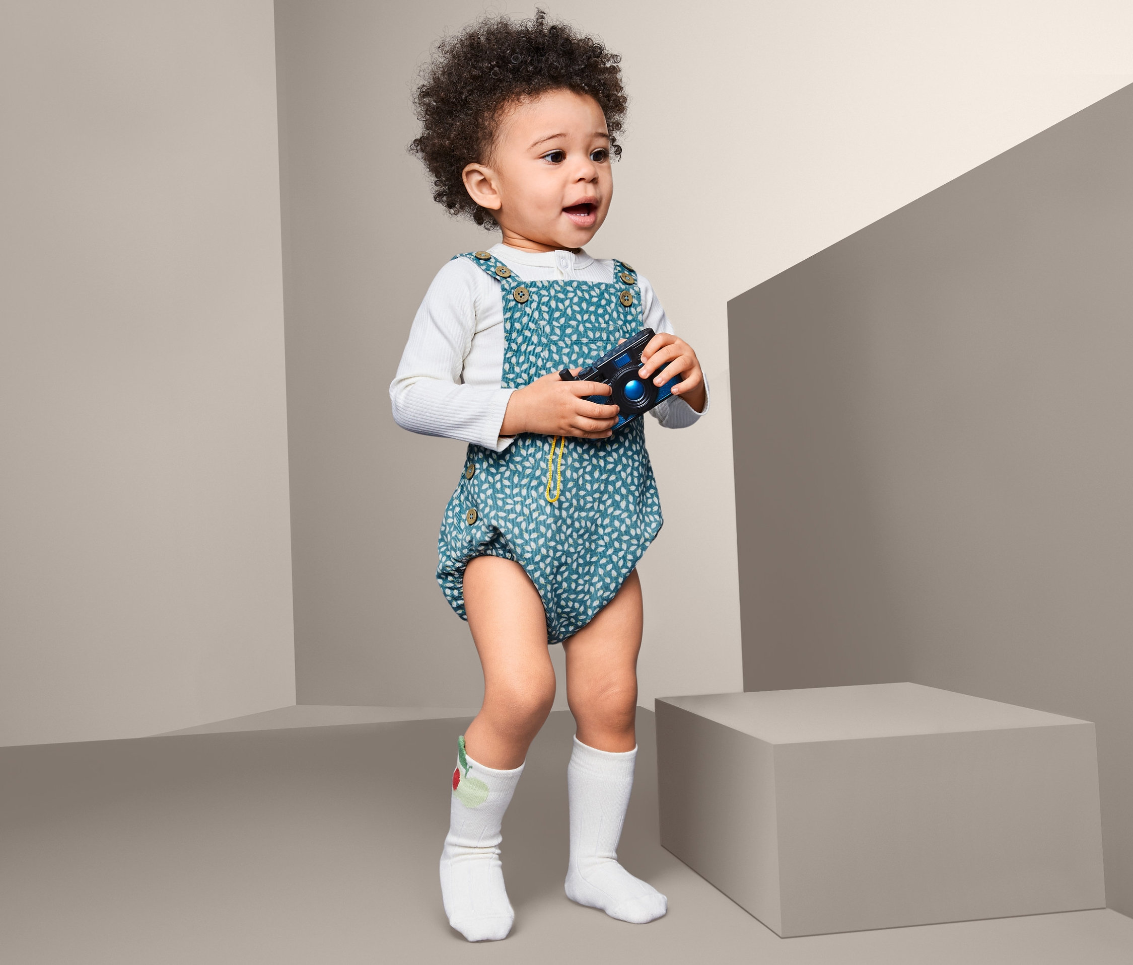 Dieťa s kučeravými vlasmi stojí a drží hračku fotoaparát. Má na sebe dupačky z jemného menčestru.