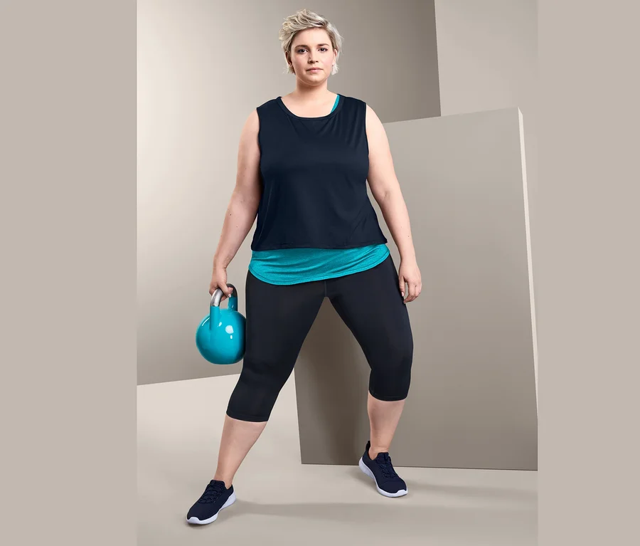 Žena drží kettlebell a má na sebe športový top 2 v 1 a trojštvrťové športové legínsy.