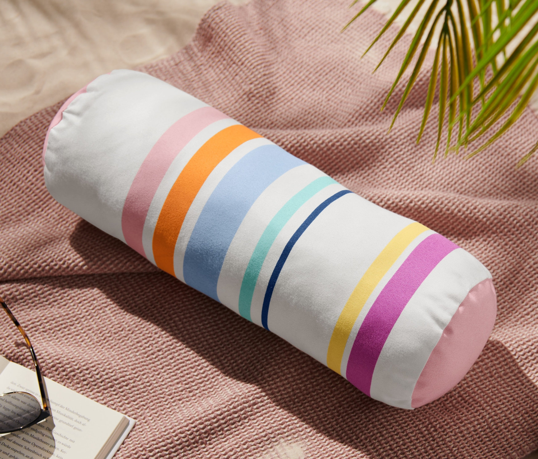 Vonkajší vankúš v tvare valca leží na ružovej deke vedľa knihy a slnečných okuliarov.
