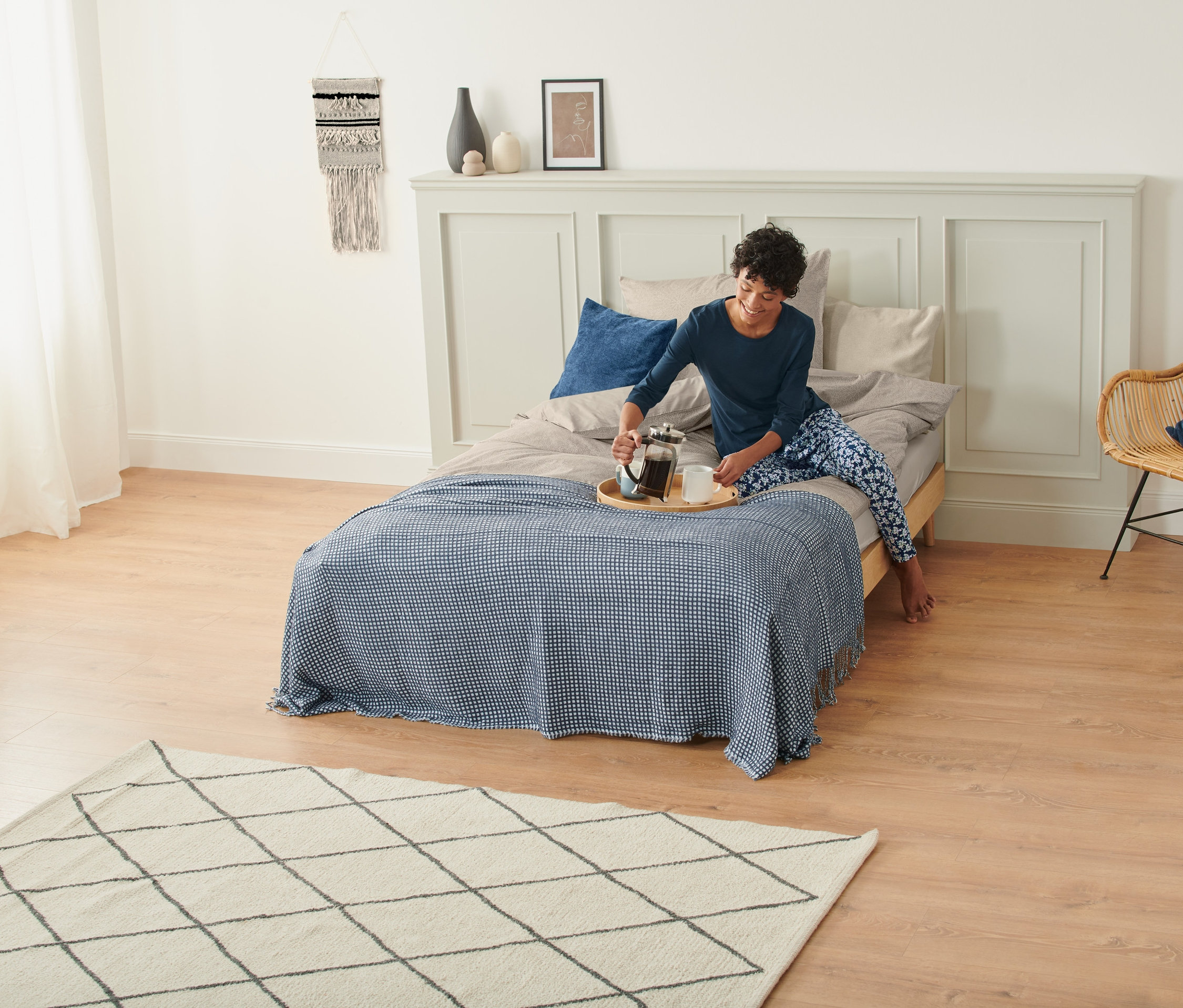 Žena v pyžame nalieva kávu z french pressu do hrnčeka na posteli s dekou a makramé kobercom na stene.