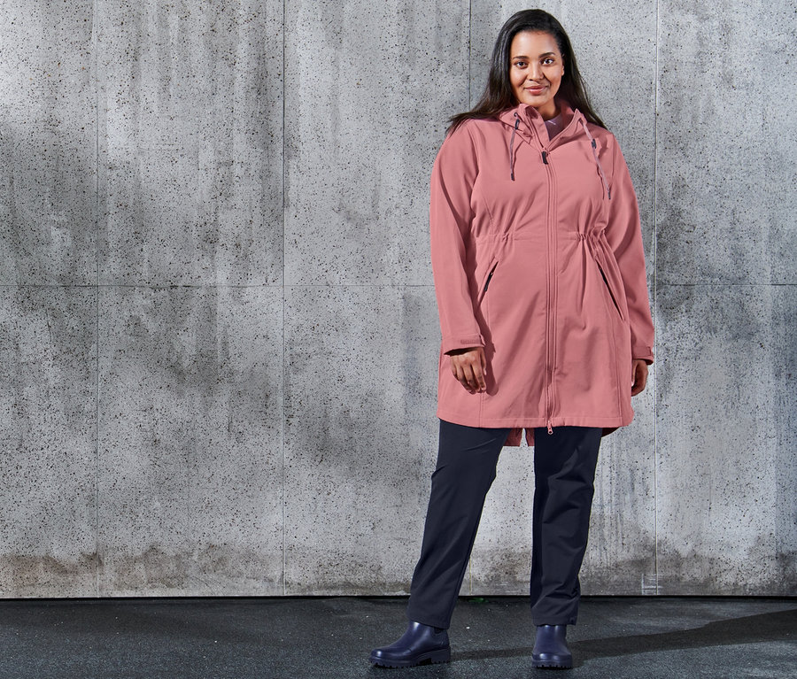Žena pózuje na pozadí betónovej steny a má na sebe softshellový kabát a softshellové tepláky tmavomodrej farby.