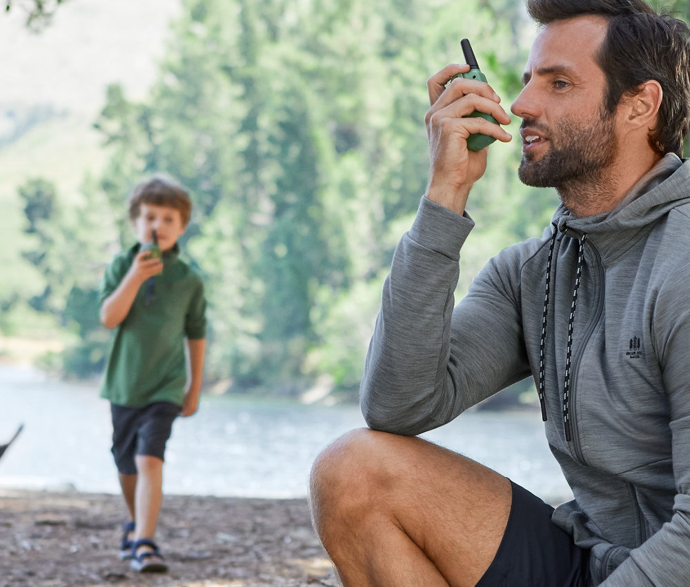 Muž sedí na kmeni a používa vysielačku. V pozadí je chlapec s vysielačkou. Obaja sú oblečení vo funkčnom oblečení.