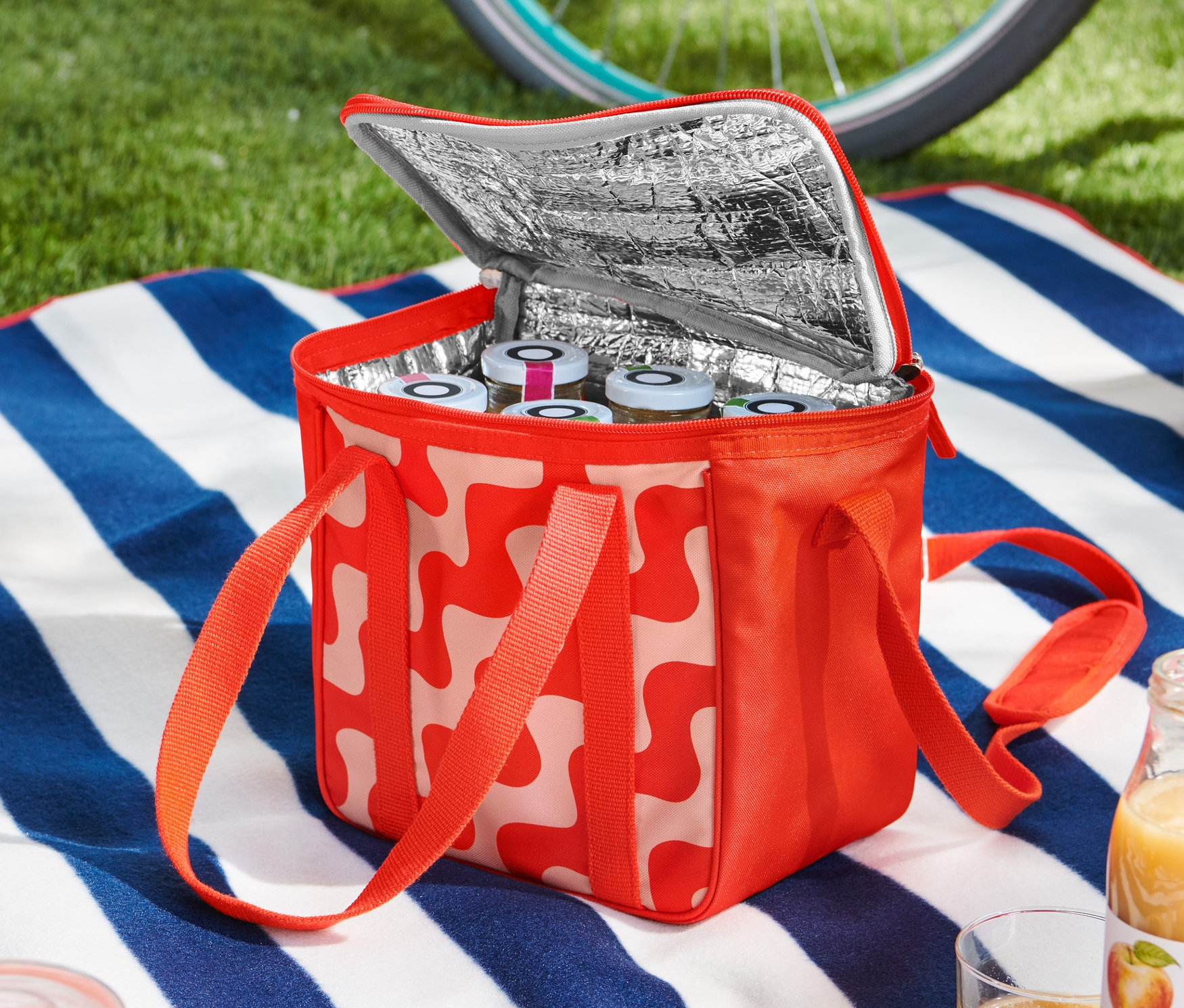 Otvorená malá chladiaca taška s červeno-bielym vzorom stojí na deke s modro-bielymi pruhmi. Vnútri sú vidieť poháre. Bicykel a helma ležia v pozadí.