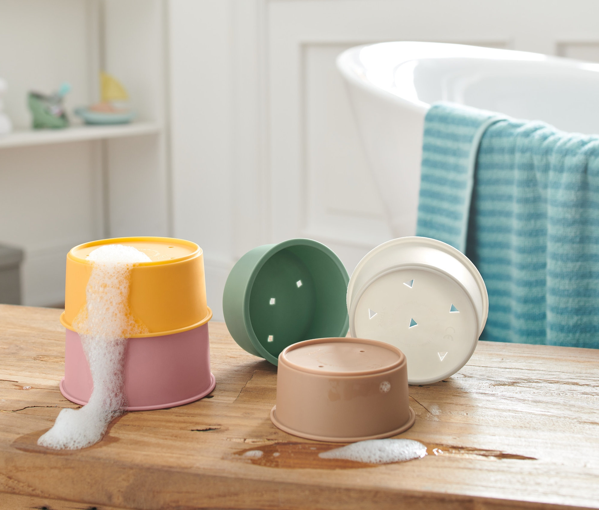 Na drevenom stole sú stohovateľné poháriky na kúpanie, dva na sebe s penou vytekajúcou z horného pohárika.