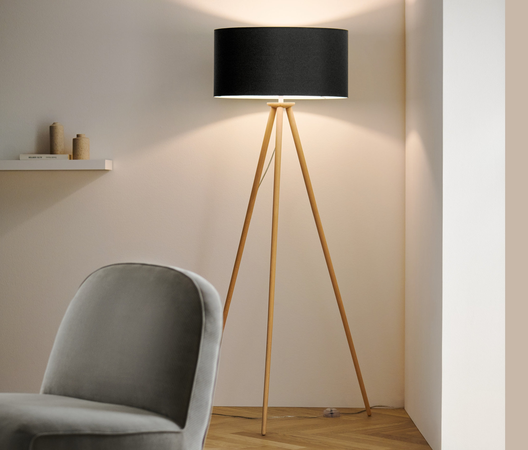 V izbe stojí drevená stojacia lampa »Trojnožka« s čiernym tienidlom a sivé kreslo.
