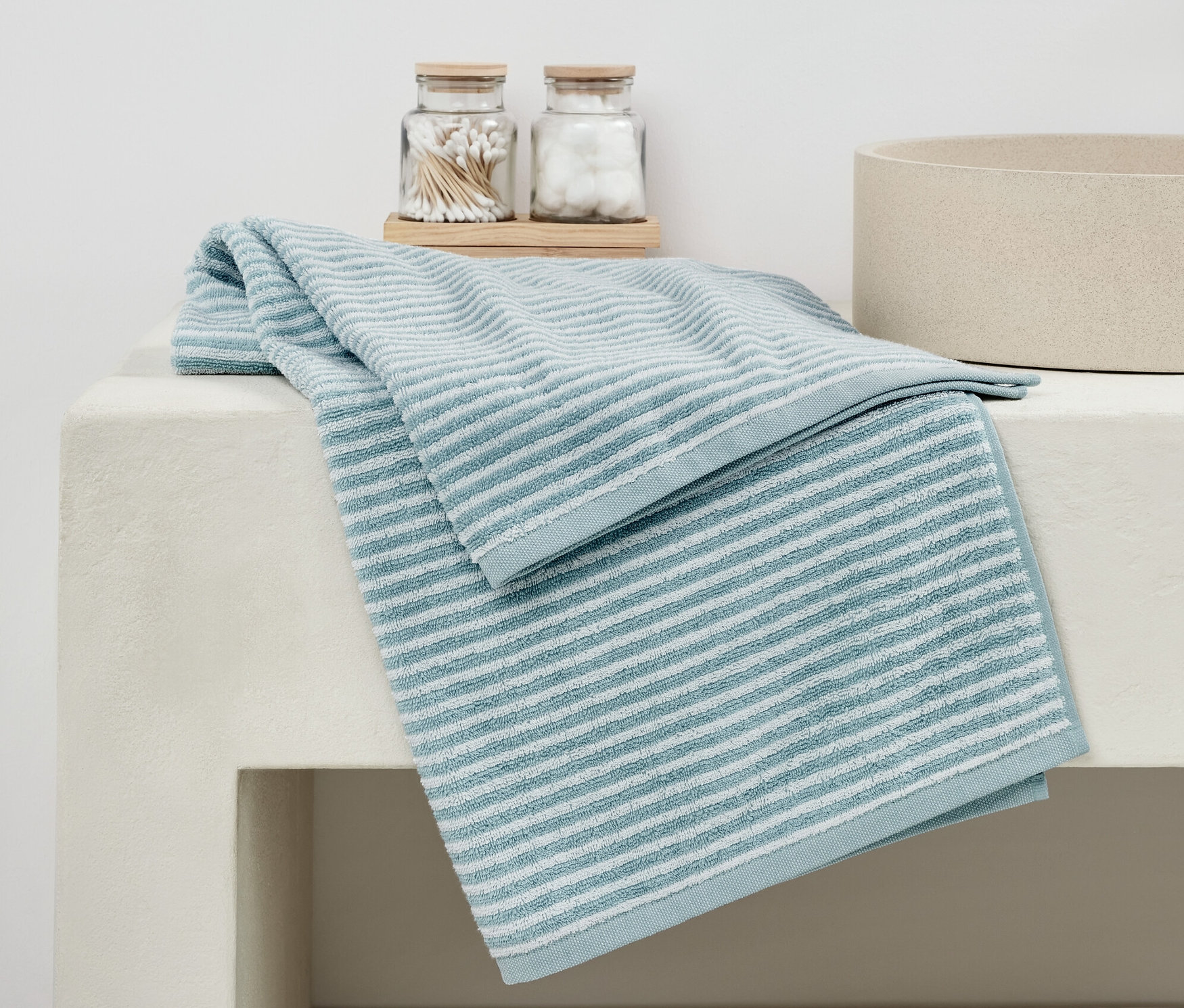 Modrá kvalitná osuška leží zložená na bielom umývadle vedľa pohárov s vatovými tyčinkami a guličkami.