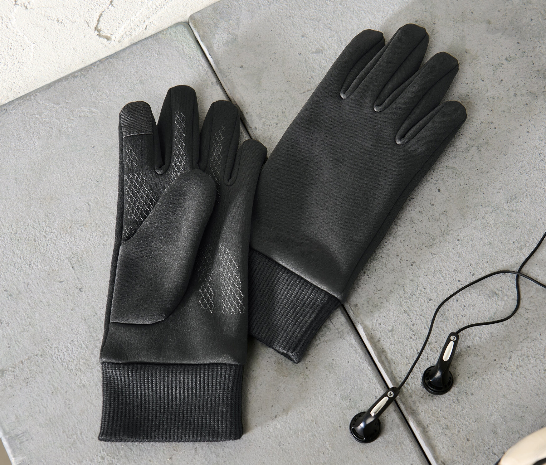 Dvoje detské vetruvzdorné termorukavice ležia na sivom povrchu vedľa slúchadiel a časti futbalovej lopty.