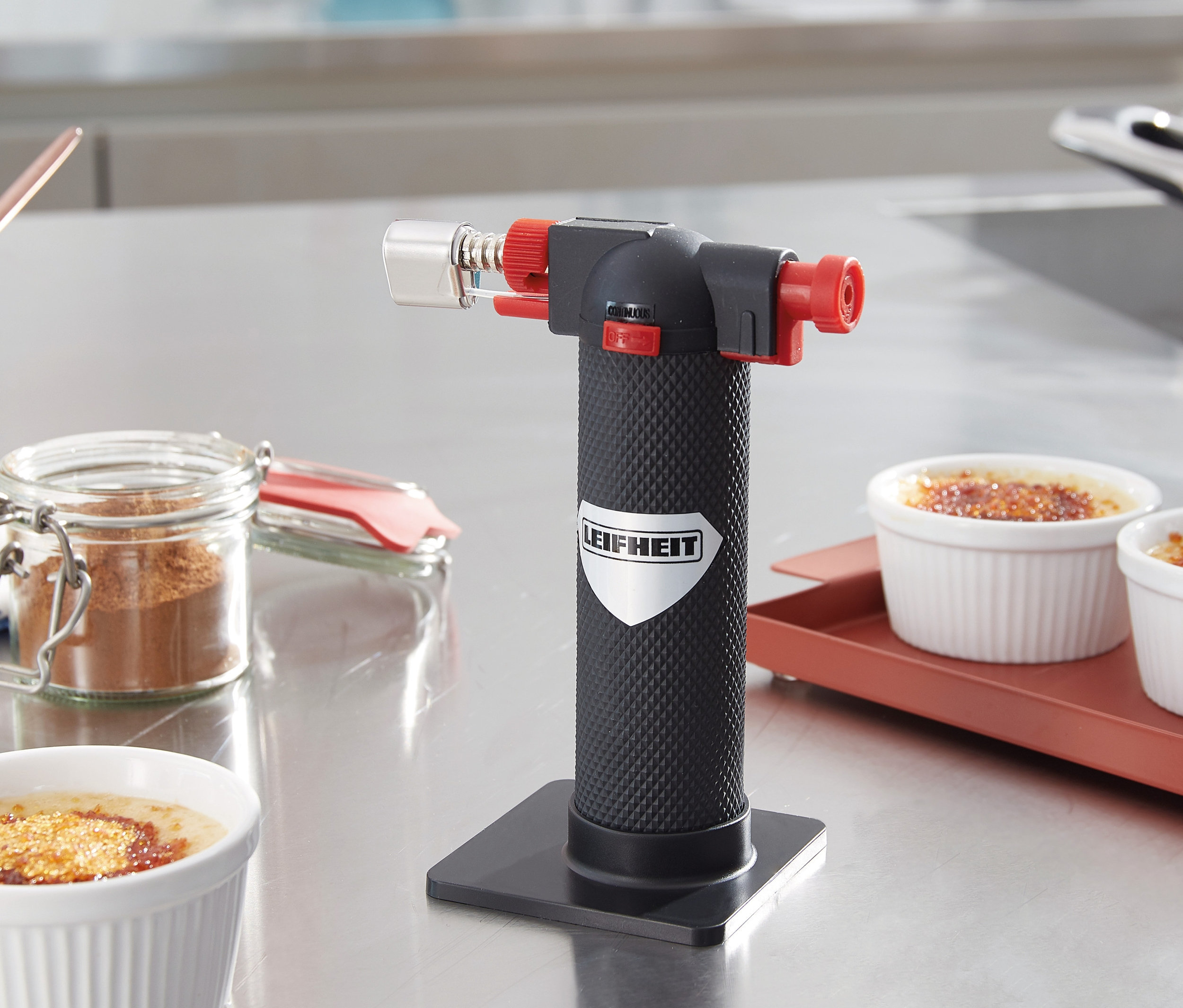 Na stole stojí kuchynská flambovacia pištoľ vedľa misiek s crème brûlée.