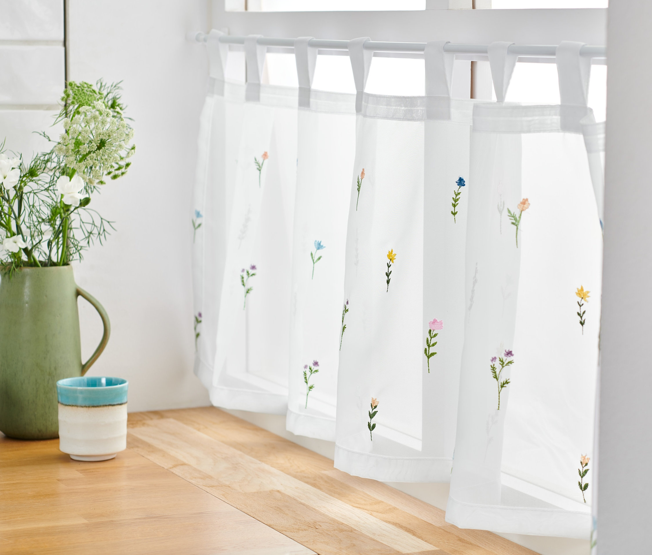 Biela krátka záclona s výšivkou kvetov visí v okne.