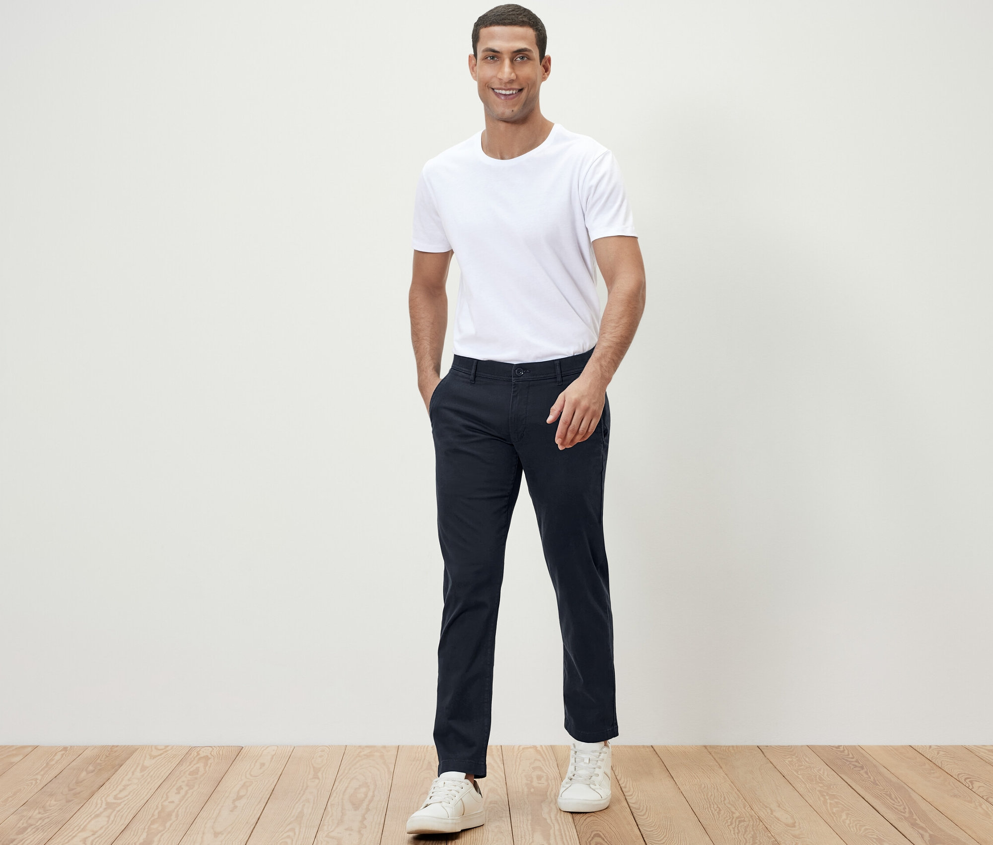 Muž pózuje do kamery v bielom tričku a tmavomodrých chino nohaviciach.