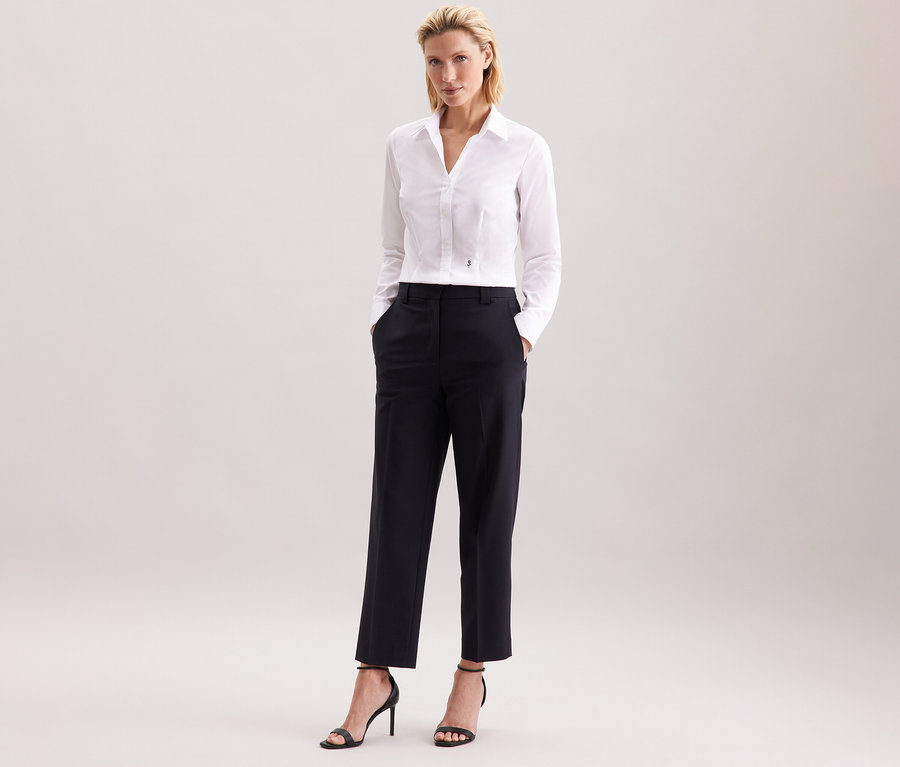 Žena stojí a má na sebe bielu blúzku Seidensticker Slim Fit non iron, čierne nohavice a čierne vysoké podpätky.