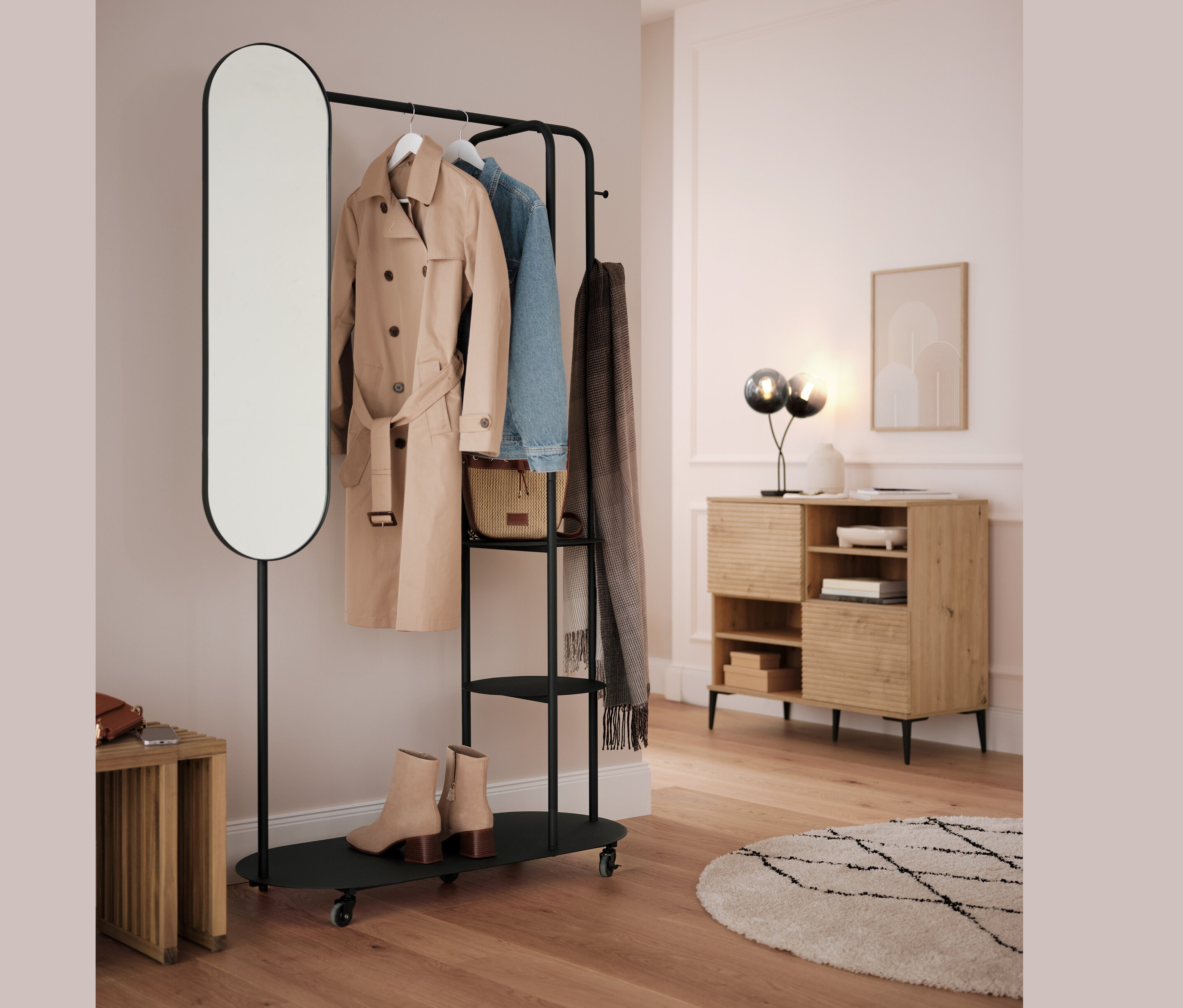Čierny vešiak na oblečenie s oválnym zrkadlom a vysúvacím stolčekom. Na tyči visí béžový kabát, modrá bunda a sivý šál.