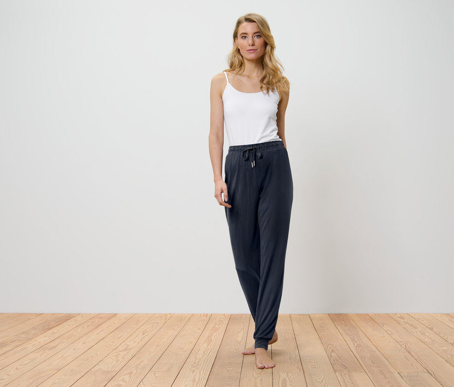 Blondínka stojí na drevenej podlahe a má na sebe biely top a tmavomodré nohavice.