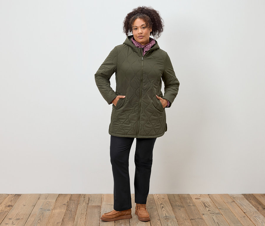 Žena stojí s rukami vo vreckách olivovozelenej prešívanej bundy s kapucňou, čiernych nohaviciach a hnedých mokasínach.
