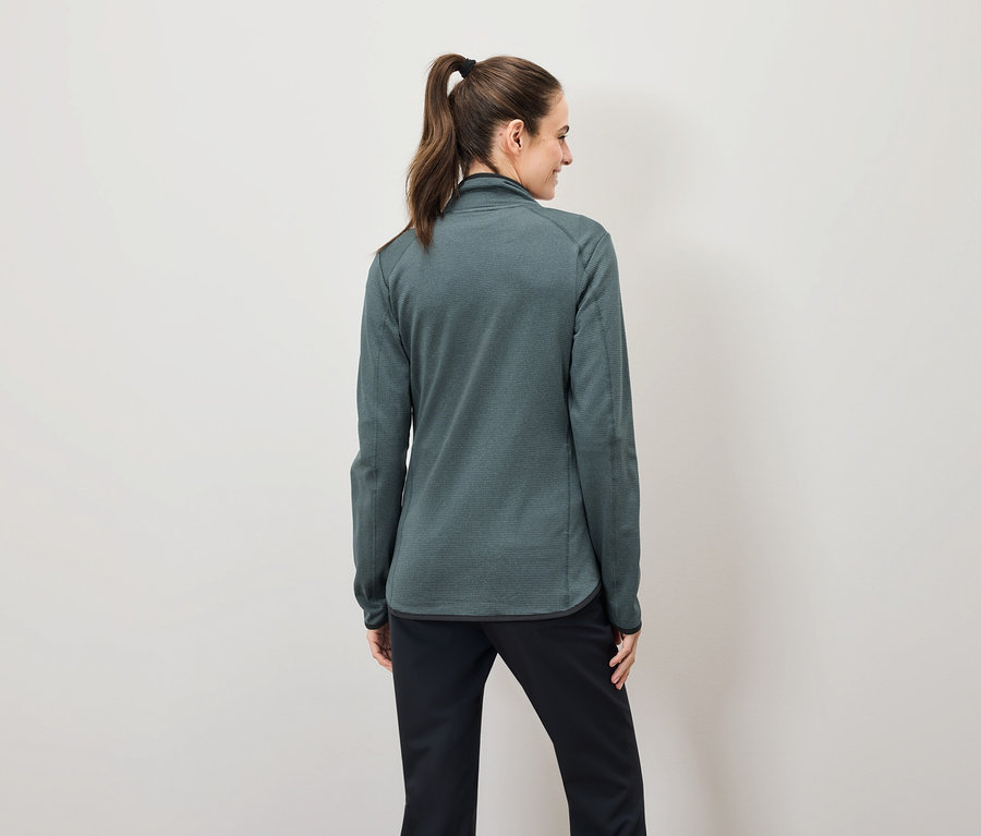 Zozadu je vidieť žena s hnedými vlasmi stiahnutými do copu, ktorá má na sebe zelenú mikinu na zips a čierne nohavice.