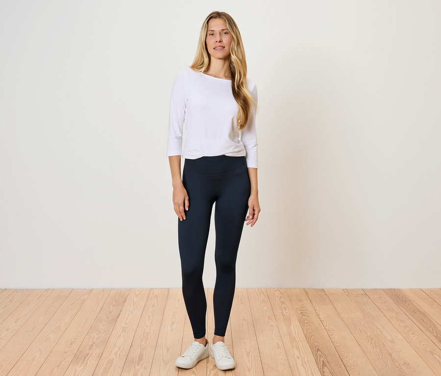 Žena stojí na drevenej podlahe a má na sebe biele tričko s dlhým rukávom a čierne legíny.