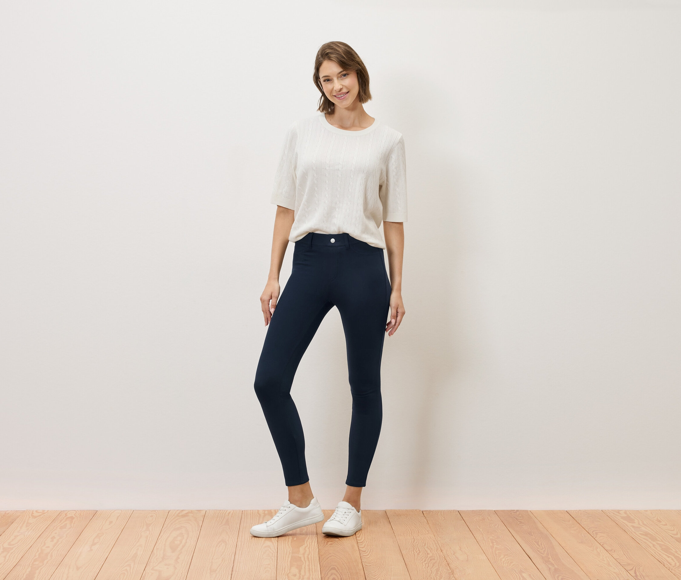 Žena pózuje v krémovom tope, tmavomodrých nohaviciach a bielych teniskách.
