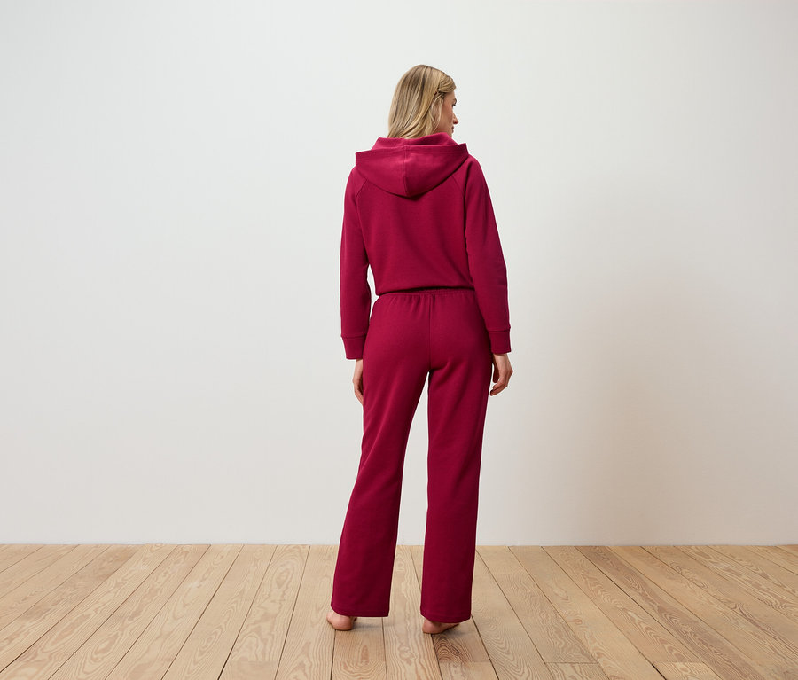 Žena stojí chrbtom, oblečená do bordovej teplákovej súpravy s kapucňou.