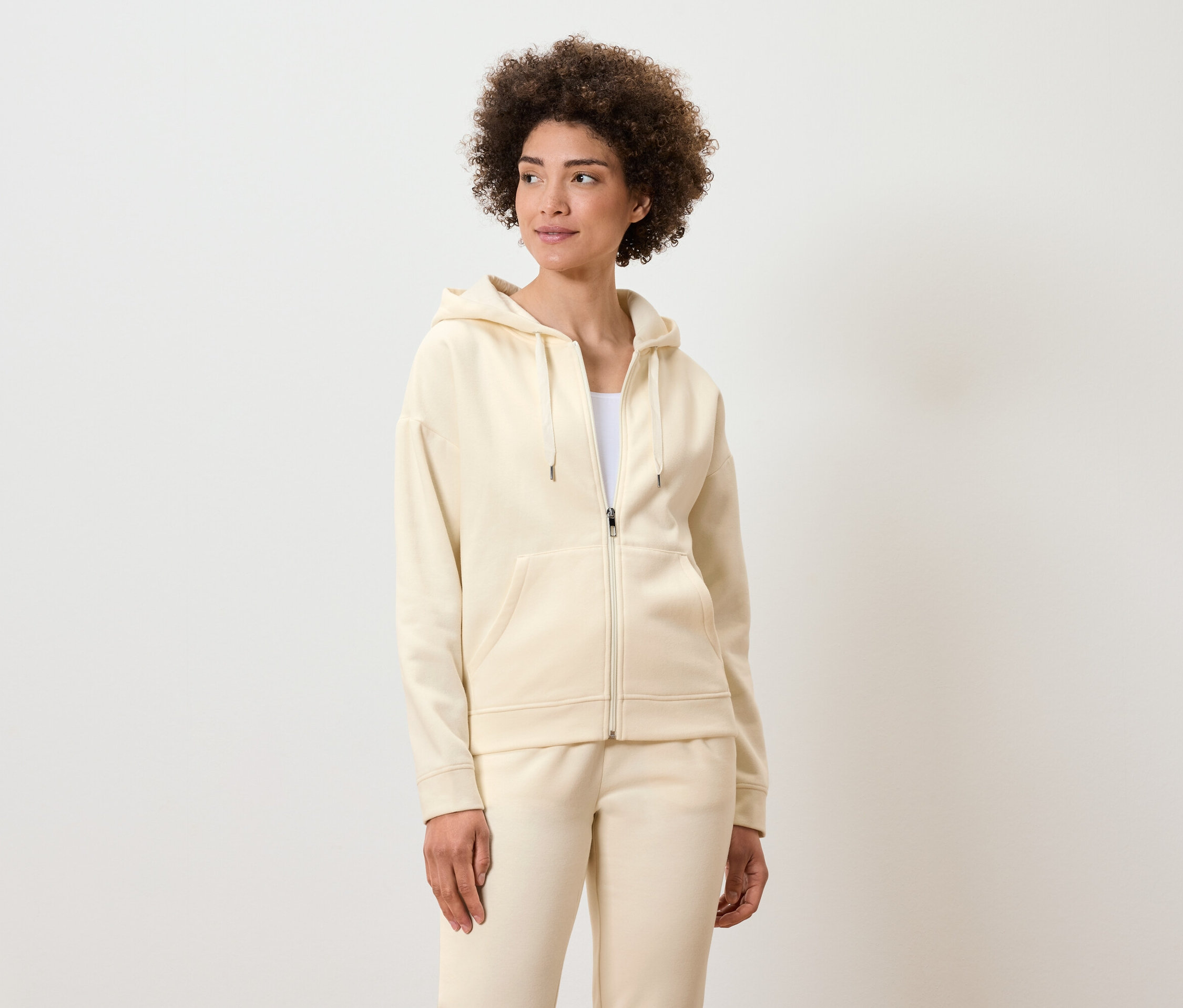 Žena s kučeravými vlasmi pózuje v krémovej mikine na zips s kapucňou a teplákoch.