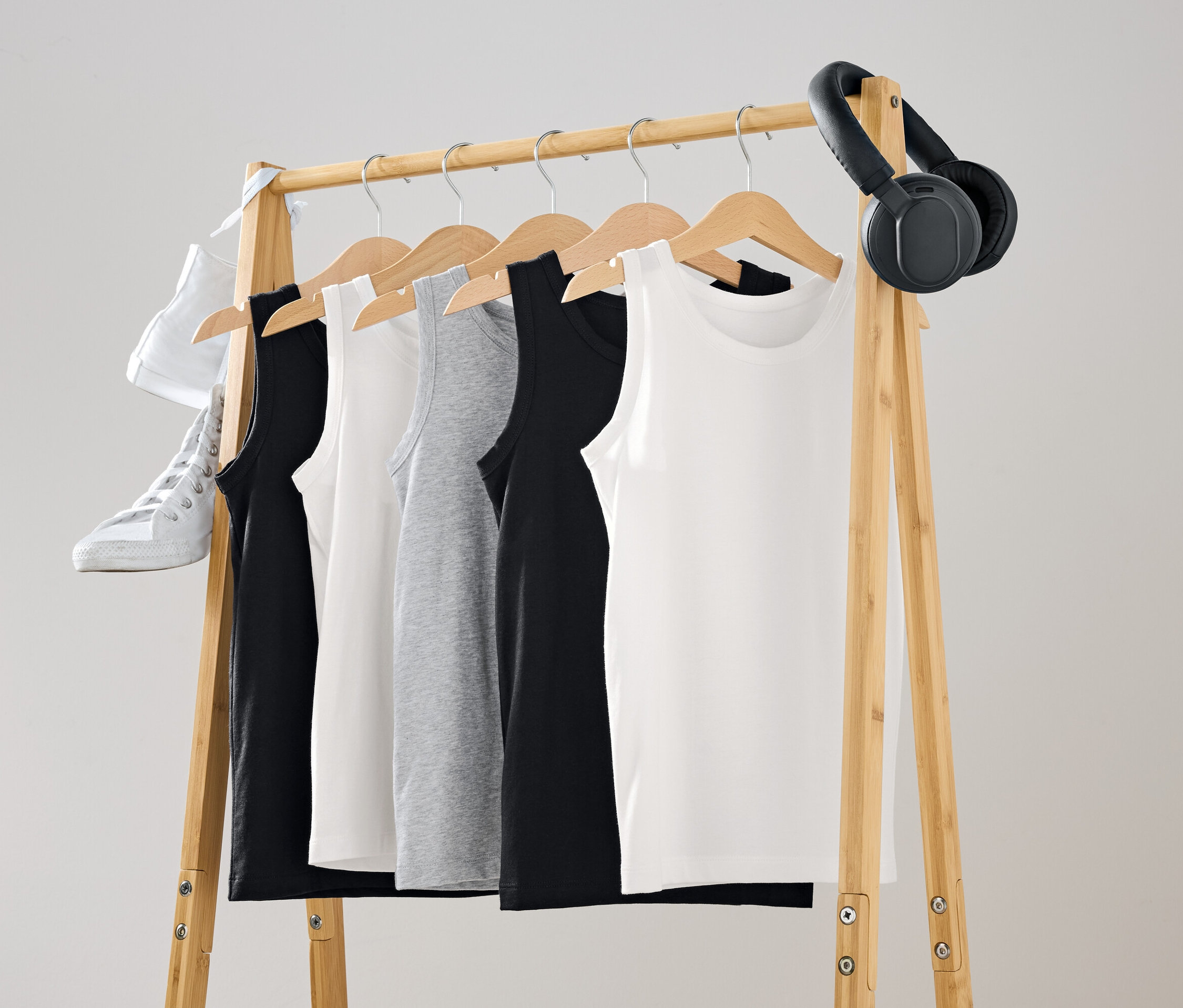 Na drevenom vešiaku visia tielka v čiernej, bielej a šedej farbe. Na vešiaku je zavesená biela topánka a čierne slúchadlá.