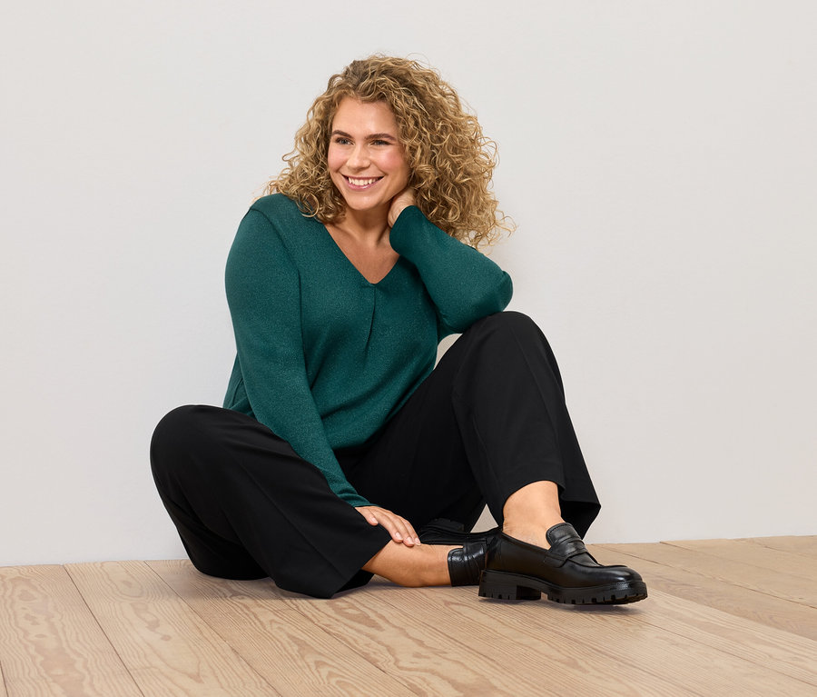 Žena sedí na podlahe v zelenom svetri a čiernych nohaviciach.