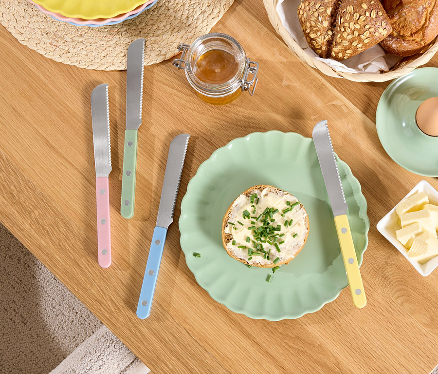 Na drevenom stole leží tanier s chlebom s maslom a bylinkami, maslo v miske, vajce v držiaku a nože s farebnými rúčkami.