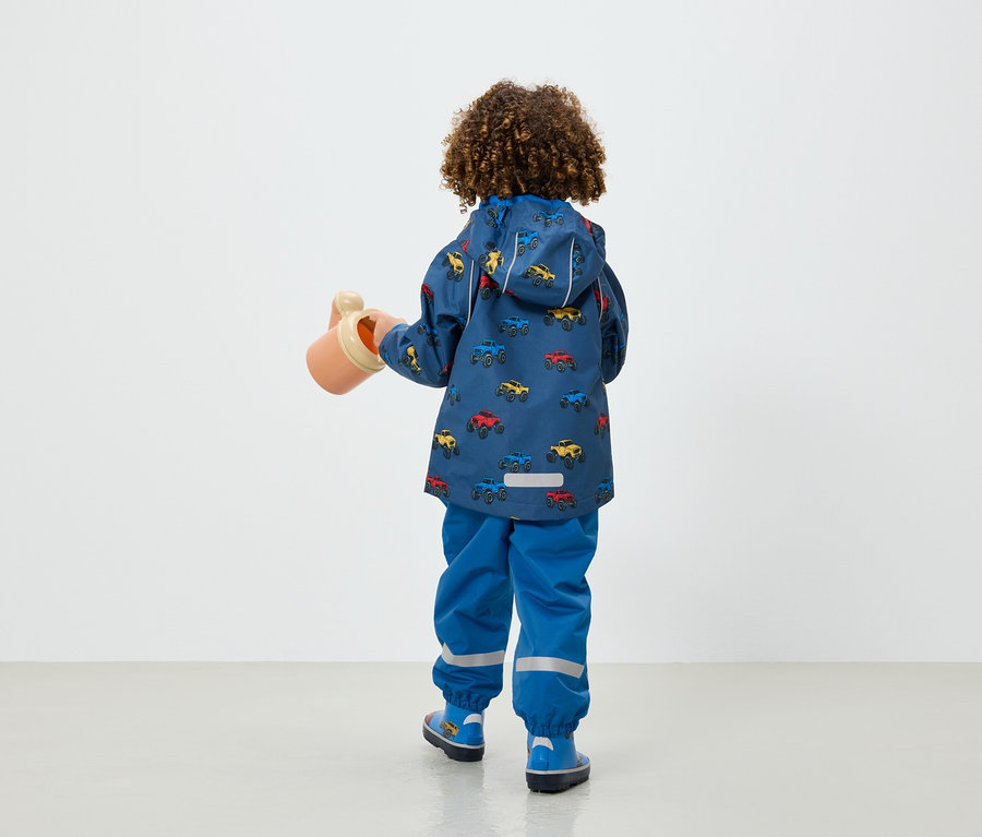 Chlapec v modrej bunde a nohaviciach s potlačou autíčok drží v ruke béžovú kanvu.