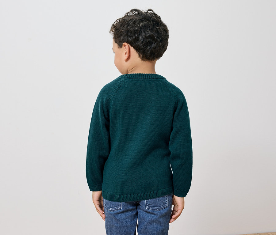 Chlapec s tmavými kučeravými vlasmi stojí chrbtom k fotoaparátu a má na sebe zelený sveter a džínsy.