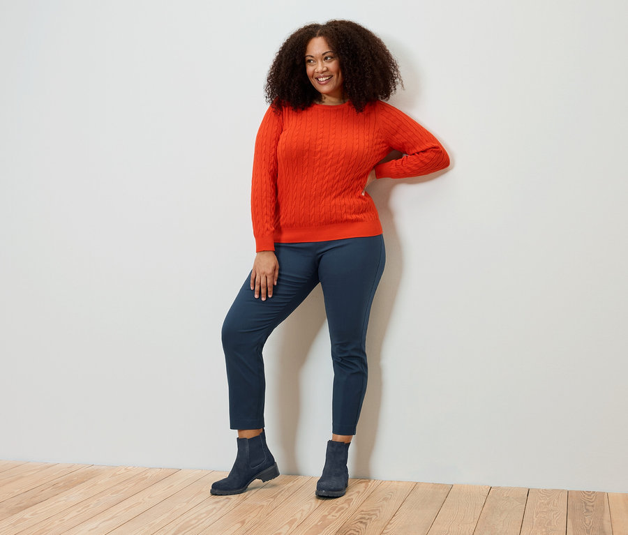 Usmievavá sa žena s kučeravými vlasmi pózuje v oranžovom svetri, modrých nohaviciach a topánkach.