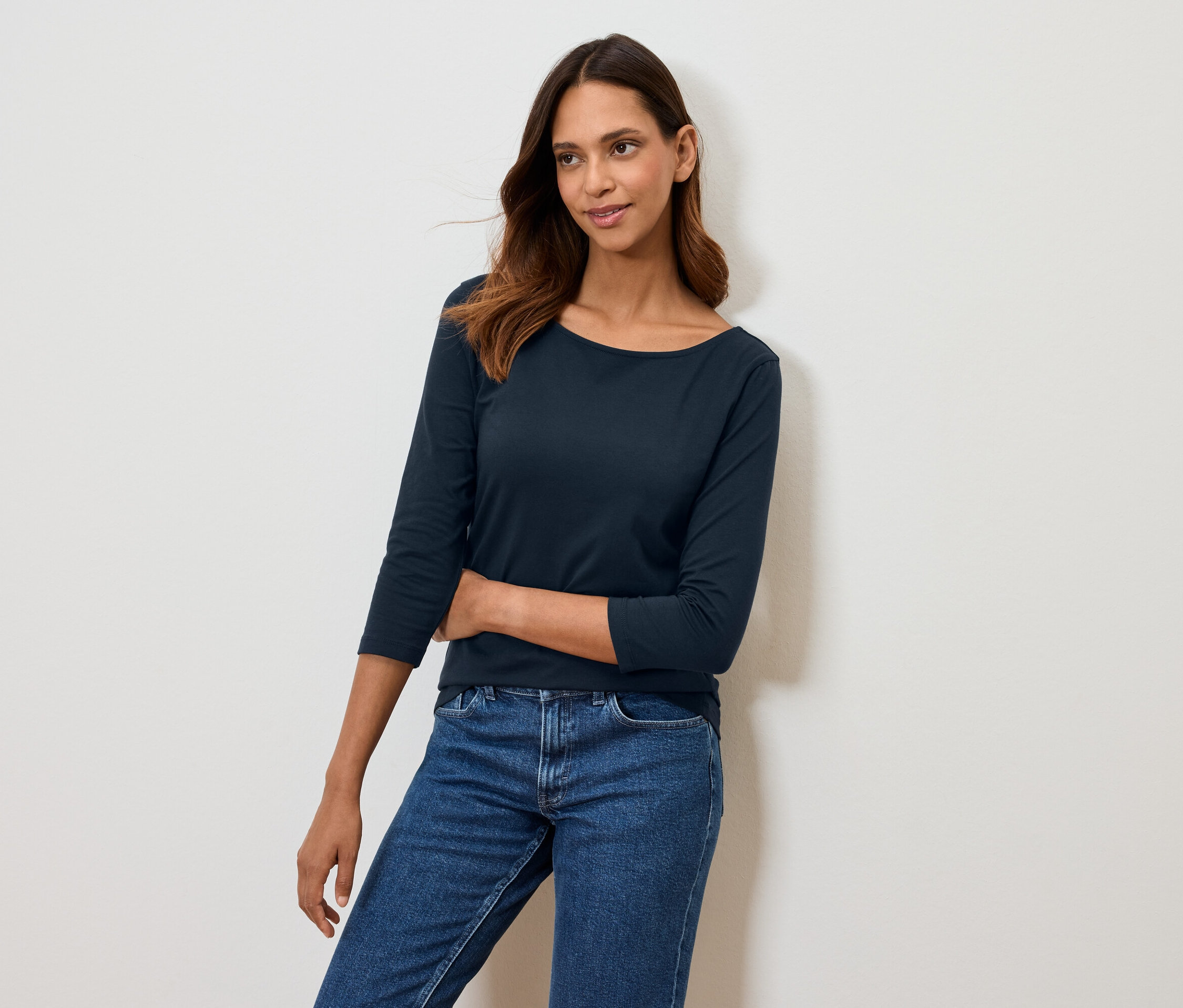 Žena pózuje v tmavomodrom tričku s trojštvrťovým rukávom a modrých džínsoch.