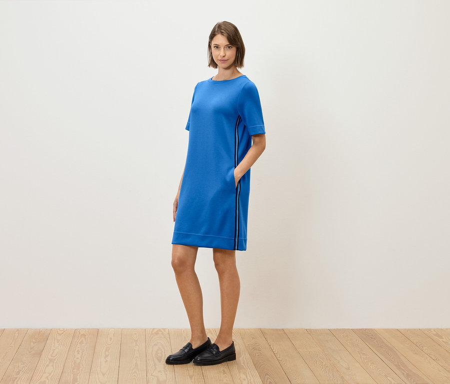 Žena stojí a pózuje v modrých šatách s krátkym rukávom a čiernych mokasínach.