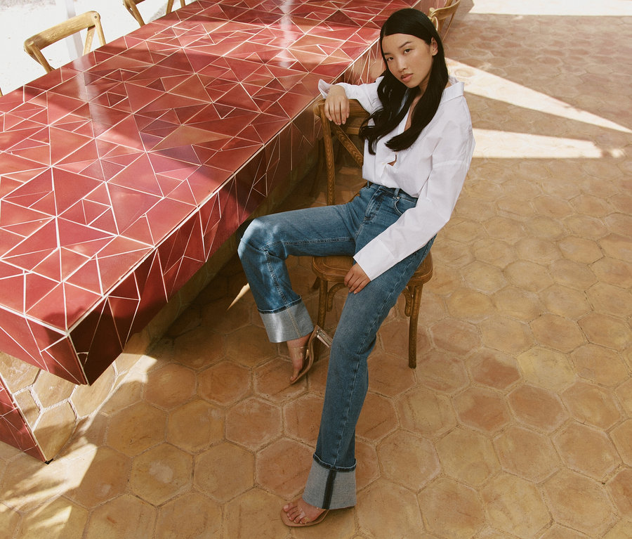 Žena sedí na stoličke pri stole a má na sebe svetlomodré džínsy Angels »Lara« rovný strih Turn Up.