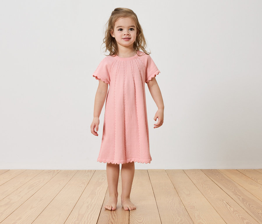 Dievča stojí bosé na drevenej podlahe a má na sebe ružové šaty.