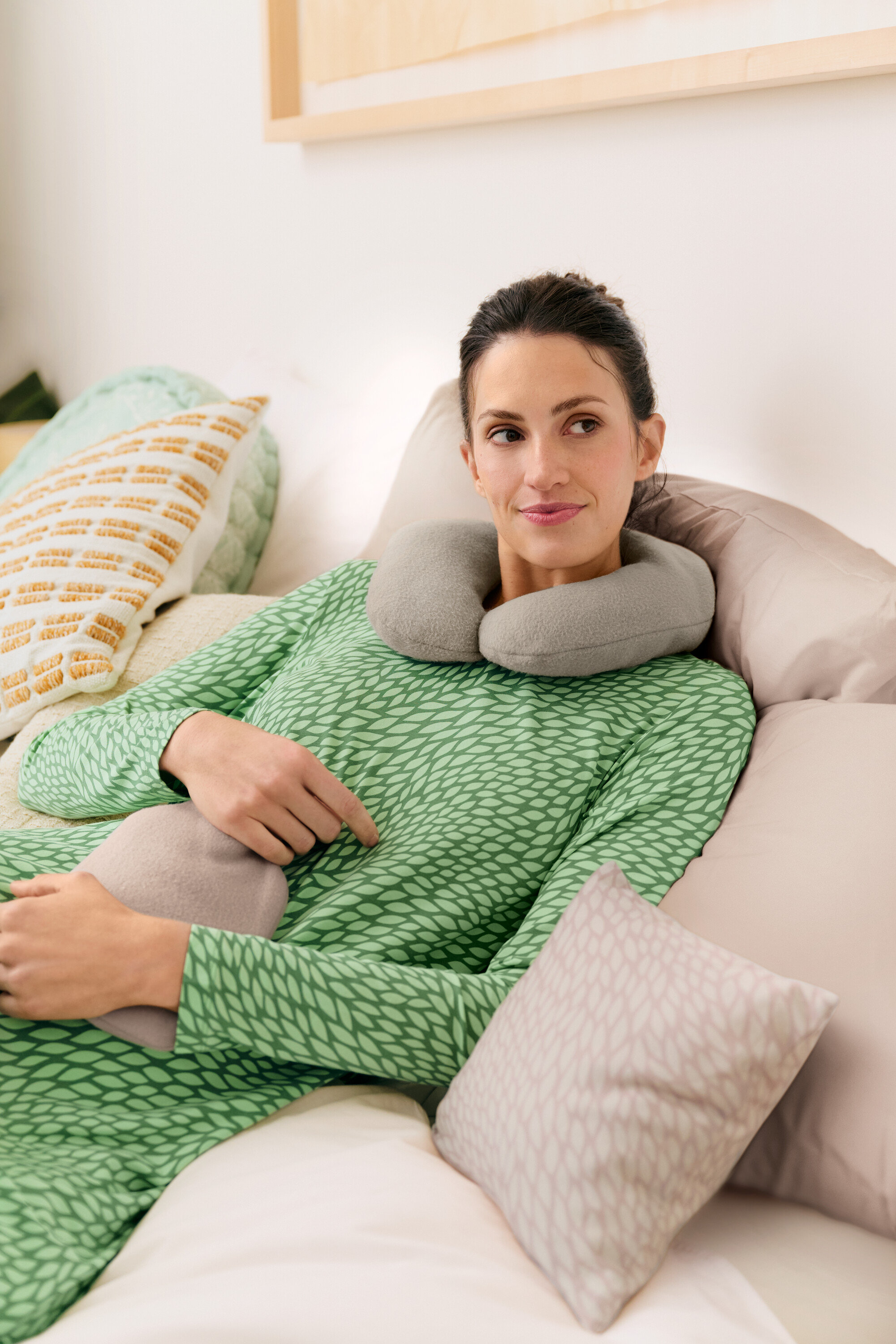 Žena leží v posteli so sivým cestovným vankúšom a termoforom, oblečená v zelenej nočnej košeli.