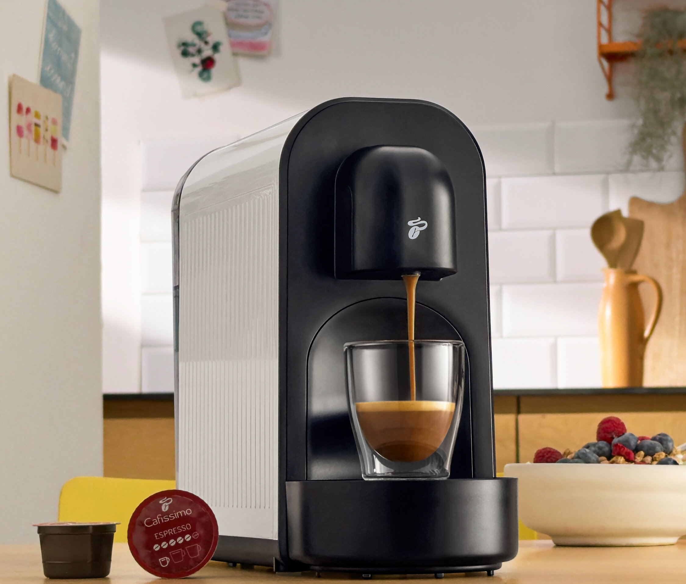 Čierny kávovar nalieva kávu do pohára. Vedľa neho ležia kávové kapsuly a miska s bobuľami.