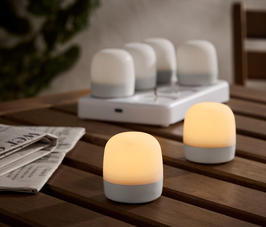 Dve osvetlené, bezdrôtové stolové lampy na drevenom stole, s nabíjacou stanicou a štyrmi ďalšími lampami v pozadí. Vedľa je noviny.