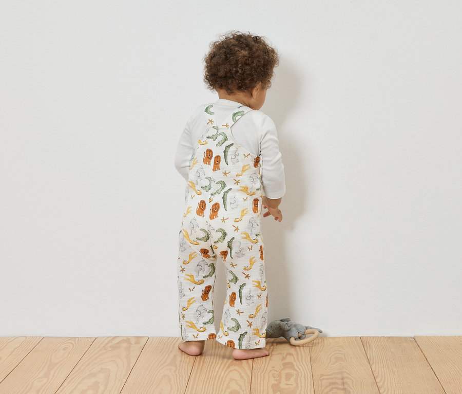 Batoľa zozadu v mušelínových nohaviciach s náprsenkou so zvieracou potlačou pred bielou stenou.