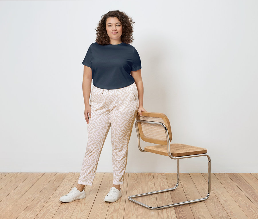 Žena s kučeravými vlasmi stojí vedľa stoličky s dreveným rámom a ratanovým sedadlom. Má na sebe tmavomodré tričko, vzorované nohavice a biele tenisky.
